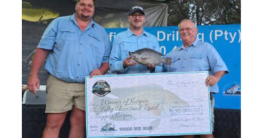 Three men wearing blue shirts stand together outdoors holding a large check for fifty thousand rand. The middle man holds a fish, smiling. It's an award ceremony.