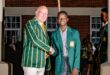 Two people wearing green blazers with gold stripes and badges shake hands, smiling. They stand in a room with brick walls and windows, exuding a celebratory atmosphere.