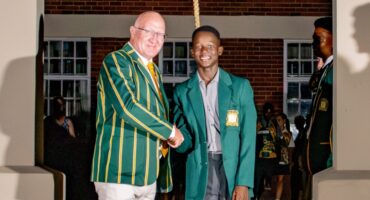 Two people wearing green blazers with gold stripes and badges shake hands, smiling. They stand in a room with brick walls and windows, exuding a celebratory atmosphere.