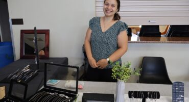 Smiling woman stands behind a table displaying assorted jewelry, including necklaces and earrings. The setting is bright and inviting, with a vase of greenery.