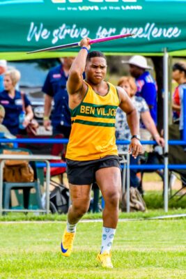 Athlete in a yellow top prepares to throw a javelin on a grassy field, with focused expression. Spectators and a green banner are in the background.