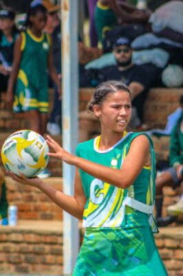 A young woman in a green netball uniform holds a ball, looking focused. Background shows spectators seated on brick steps, some players in similar outfits.