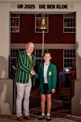 A man and a young student, both in green blazers, shake hands under a school bell. The background shows a brick building and people watching. Text reads "UR 2025 DIE BEN KLOK."