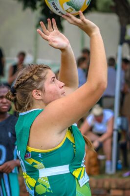 A netball player in a green and yellow uniform is focused and poised to take a shot, with hands raised, in an outdoor setting. Spectators are blurred behind her.