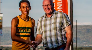 A young athlete in a yellow jersey shakes hands with a man in a plaid shirt outdoors. They smile against a backdrop of hills and a red banner.