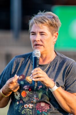 A person with short hair speaks into a microphone while wearing a t-shirt with colorful flowers. The background is blurred, featuring a green sign.