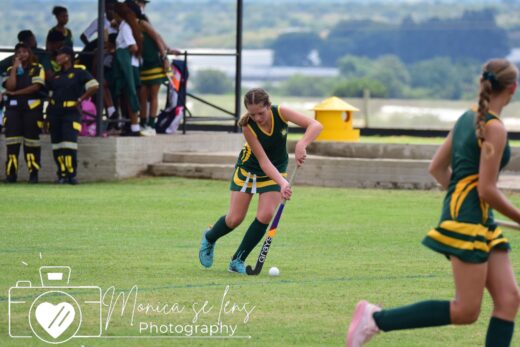 Teen girl in green and yellow uniform plays field hockey, focused on the ball. Teammates and spectators watch, creating an active sports scene.