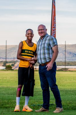 A young athlete in a yellow and black sports uniform stands with a man in a plaid shirt on a grassy field, both smiling warmly. Mountains and a flagpole are in the background.