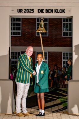 A man and a young woman in green blazers stand smiling, shaking hands under a gold bell in an archway. Background shows a brick building and students.