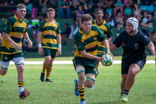 Rugby player in green and yellow uniform runs with a ball, pursued by an opponent in black. Three teammates follow on a grassy field. Competitive atmosphere.