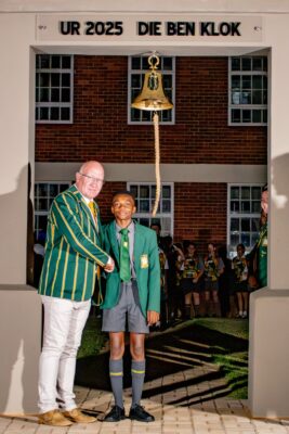 A man and a boy in striped blazers shake hands under a bell with "UR 2025 DIE BEN KLOK" above. Students in similar attire stand in the background.