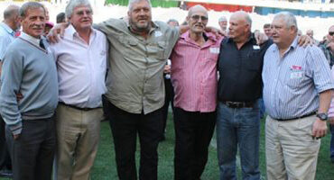 A group of six men standing together with arms around each other, smiling, in a lively outdoor setting.