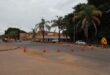 Street scene with road construction under cloudy skies. Workers in orange gear are near traffic cones, while palm trees and a strip mall line the street.