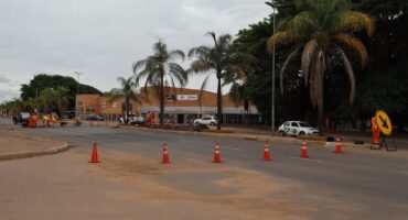 Street scene with road construction under cloudy skies. Workers in orange gear are near traffic cones, while palm trees and a strip mall line the street.