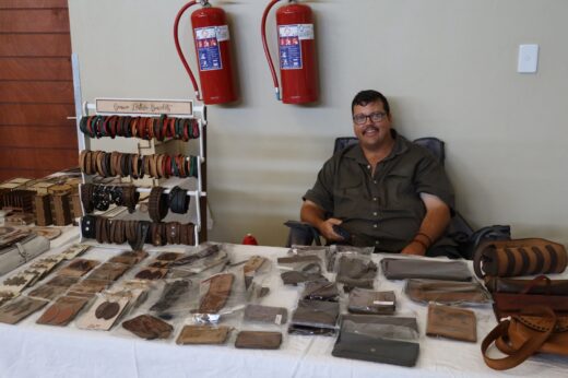 Man smiling at a market stall displaying handmade leather goods—wallets, belts, and bracelets. The setting is informal, with a relaxed atmosphere.