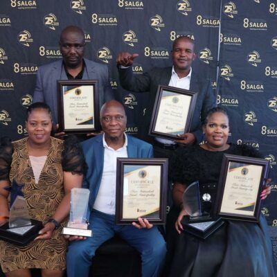 A group of six individuals posing with awards and certificates against a branded backdrop, celebrating achievements in local government.
