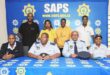 A group photo with seven people, four in police uniforms, sitting at a table with a "SAPS" blue banner in the background, conveying teamwork.