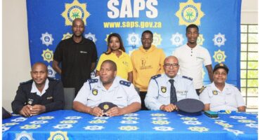 A group photo with seven people, four in police uniforms, sitting at a table with a "SAPS" blue banner in the background, conveying teamwork.