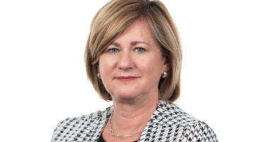 A woman with short, light brown hair, wearing a black and white houndstooth jacket and pearl earrings. She has a calm, confident expression.
