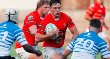 The image shows two rugby players in red jerseys running with a ball, facing opponents in blue and white. The scene conveys intensity and competition.