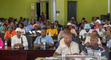 A large group of people attentively participates in a community meeting in a green-walled room. Bottled water and documents are visible on tables.