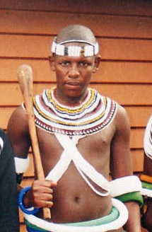 Young person wearing traditional beaded attire and headband, holding a wooden staff. The background features wooden paneling. The expression is serious.