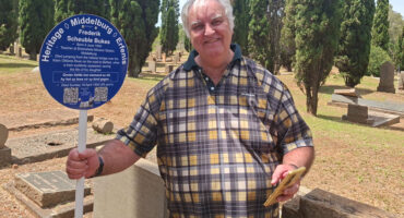 A man holding a sign in a cemetery, wearing a plaid shirt and smiling at the camera.