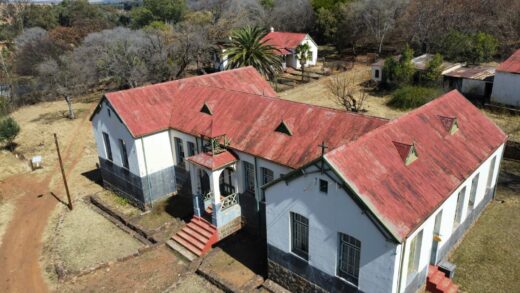 Aerial view of an old building with a red roof and white walls, surrounded by trees and grassland.