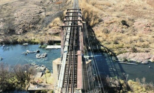 A railway bridge spans over a narrow river with rocky areas and grassy banks on either side.