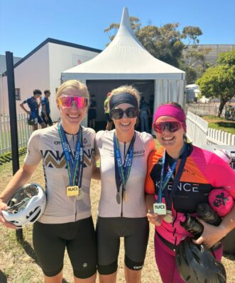 Three women in cycling gear smiling and posing together for a photo outdoors.