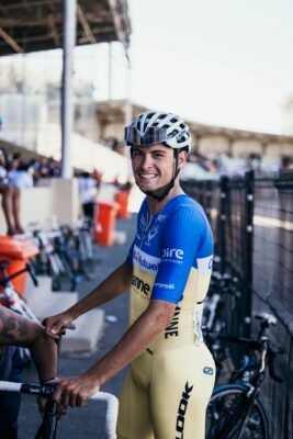 A cyclist in a yellow and blue uniform and helmet stands beside a bicycle, smiling at the camera near a fenced area at an outdoor event.