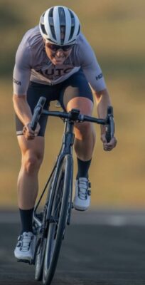 A cyclist wearing a helmet, sunglasses, and a cycling kit rides a road bike on an outdoor track