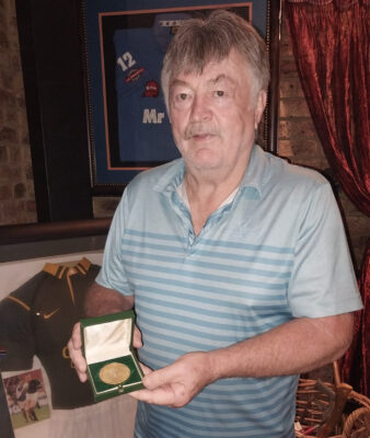 An older man proudly holds a medal in front of a framed picture, showcasing a moment of achievement and reflection.