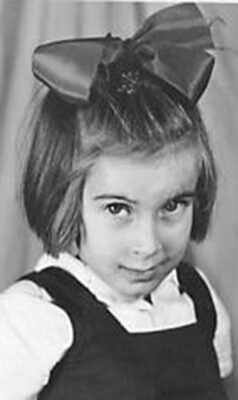A young girl with a large bow in her hair, wearing a dark dress and white shirt, is posing for a portrait.