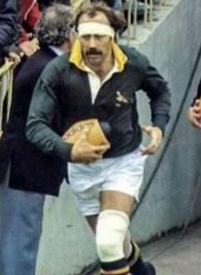 A rugby player in a green and gold jersey runs with a ball, preparing to enter the field, surrounded by spectators.