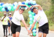 Two marathon runners in white shirts form heart shapes with their arms under a colorful balloon arch. Other runners and volunteers in the background create an upbeat and festive atmosphere.
