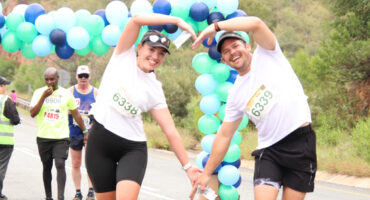 Two marathon runners in white shirts form heart shapes with their arms under a colorful balloon arch. Other runners and volunteers in the background create an upbeat and festive atmosphere.