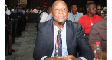 A man in a suit sits attentively at a conference, surrounded by attendees. The atmosphere is focused, with dim lighting and banners in the background.