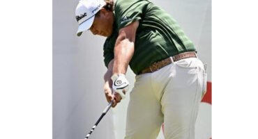 Golfer in a green shirt and beige pants swings a club on a sunny day, wearing a white cap and glove, conveying focus and determination.
