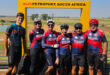 A group of six cyclists in colorful jerseys and helmets stand smiling under a yellow frame labeled "Alzu Petroport, South Africa," with a vast, sunny landscape in the background.