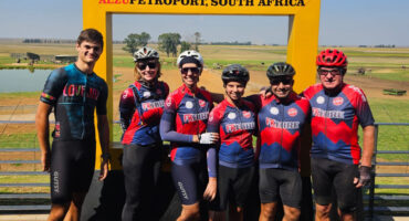 A group of six cyclists in colorful jerseys and helmets stand smiling under a yellow frame labeled "Alzu Petroport, South Africa," with a vast, sunny landscape in the background.
