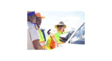 Two traffic officers in bright vests and hats converse with a driver through a car window. The scene is outdoors on a sunny day.
