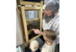 A man with a beard shows two young boys a wooden-framed glass case filled with bees. The boys listen attentively, creating an educational tone.
