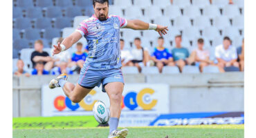 A rugby player in a gray and pink uniform prepares to kick a ball on a grassy field. Spectators are seated in the background with focused expressions.