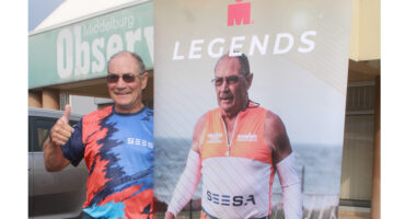 A man in colorful sportswear stands next to a banner displaying an athlete with "LEGENDS" text. He's smiling and giving a thumbs-up, exuding pride and enthusiasm.