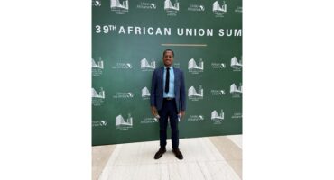 Man in a blue suit and tie stands smiling against a green backdrop reading "39th African Union Summit," conveying professionalism and positivity.