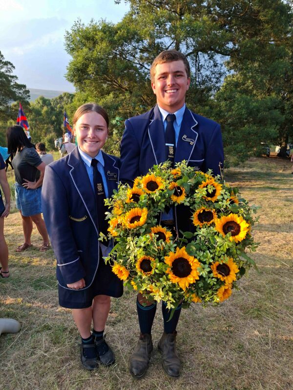 Two students in blue uniforms stand outdoors, smiling. They hold a wreath of vibrant sunflowers. Flags and trees are visible in the sunny background.