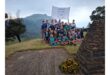 A group of young people, smiling, gather on a grassy hilltop with a scenic mountain view. They hold a flag and sit near a stone monument adorned with sunflowers.