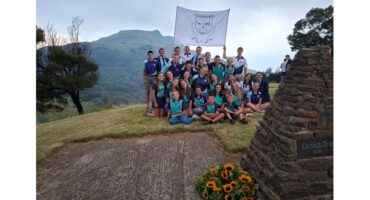 A group of young people, smiling, gather on a grassy hilltop with a scenic mountain view. They hold a flag and sit near a stone monument adorned with sunflowers.