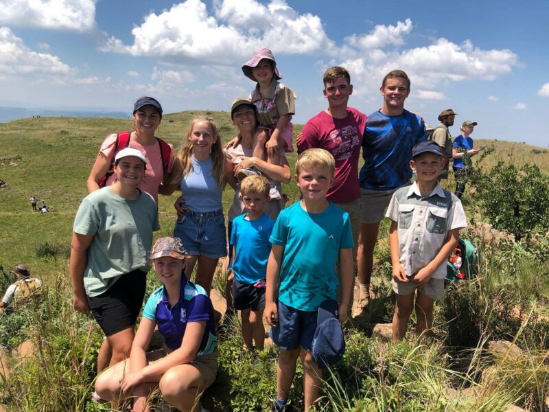 A group of smiling people poses outdoors on a grassy hill under a sunny, blue sky with clouds. The mood is cheerful and relaxed, suggesting a day of hiking or outdoor activities.